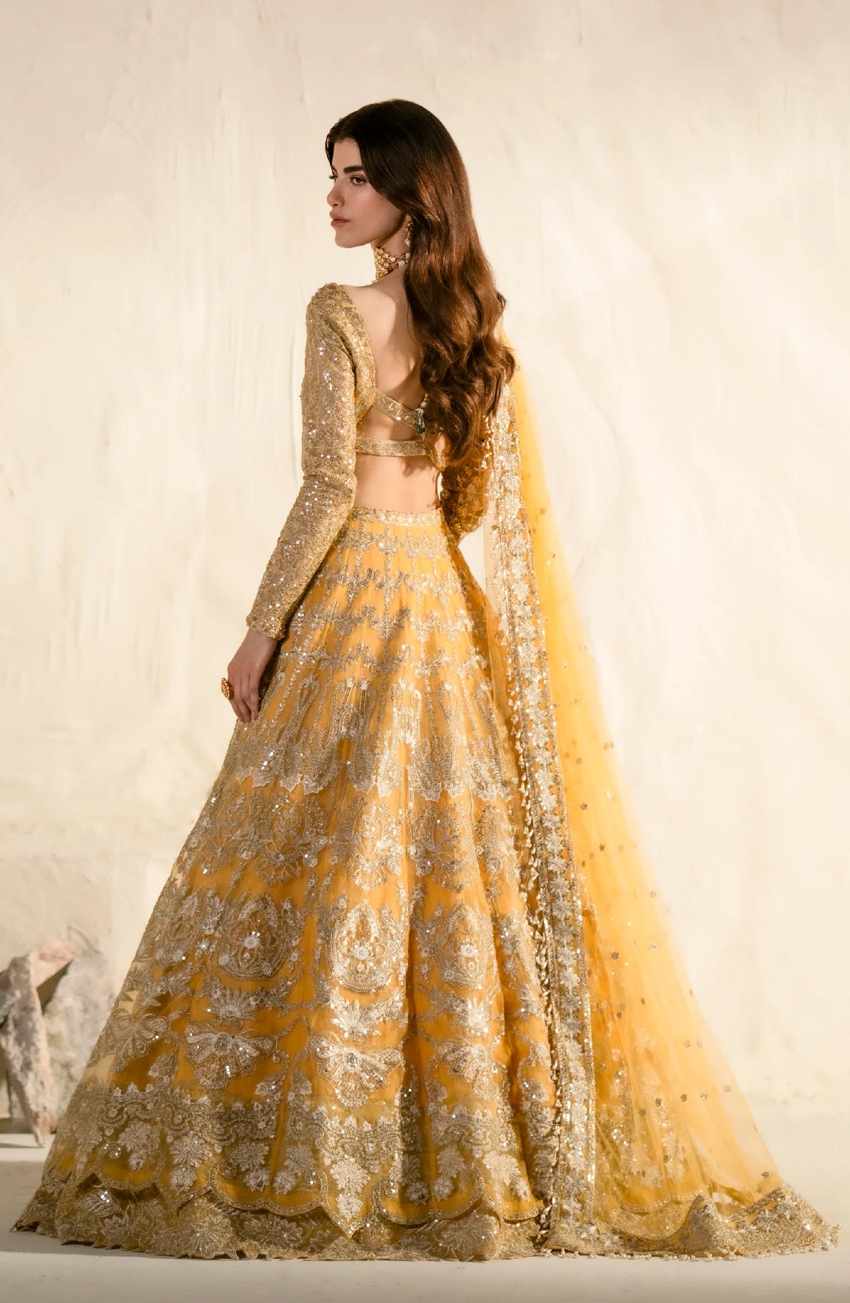 Back view of ornate golden lehenga set displaying intricate embellishments and flowing dupatta with traditional Pakistani/Indian formal wear design