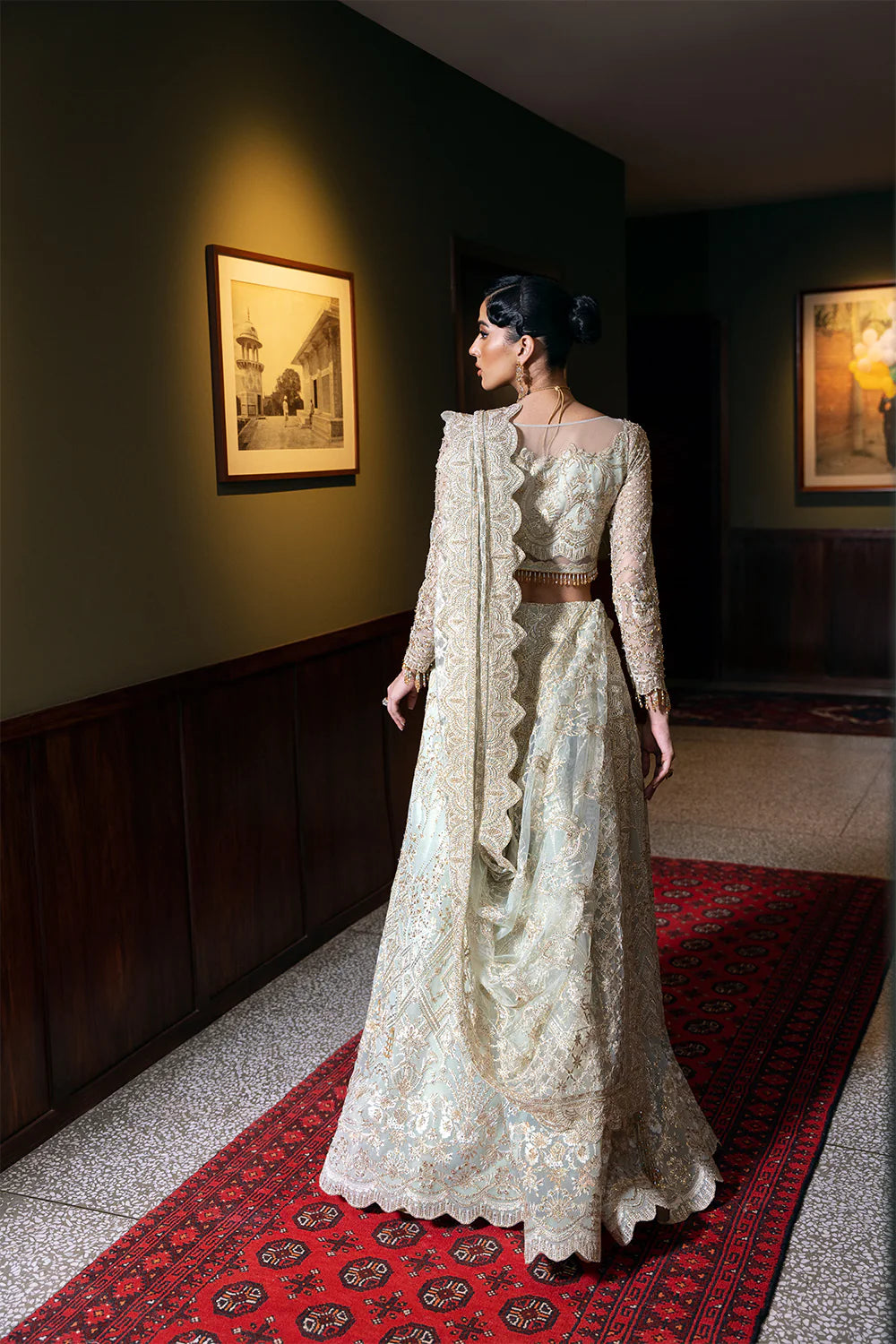 Back view of Pakistani bridal dress showing full-length silhouette with intricate embroidered details, flowing dupatta, and traditional bridal styling on decorative carpet.