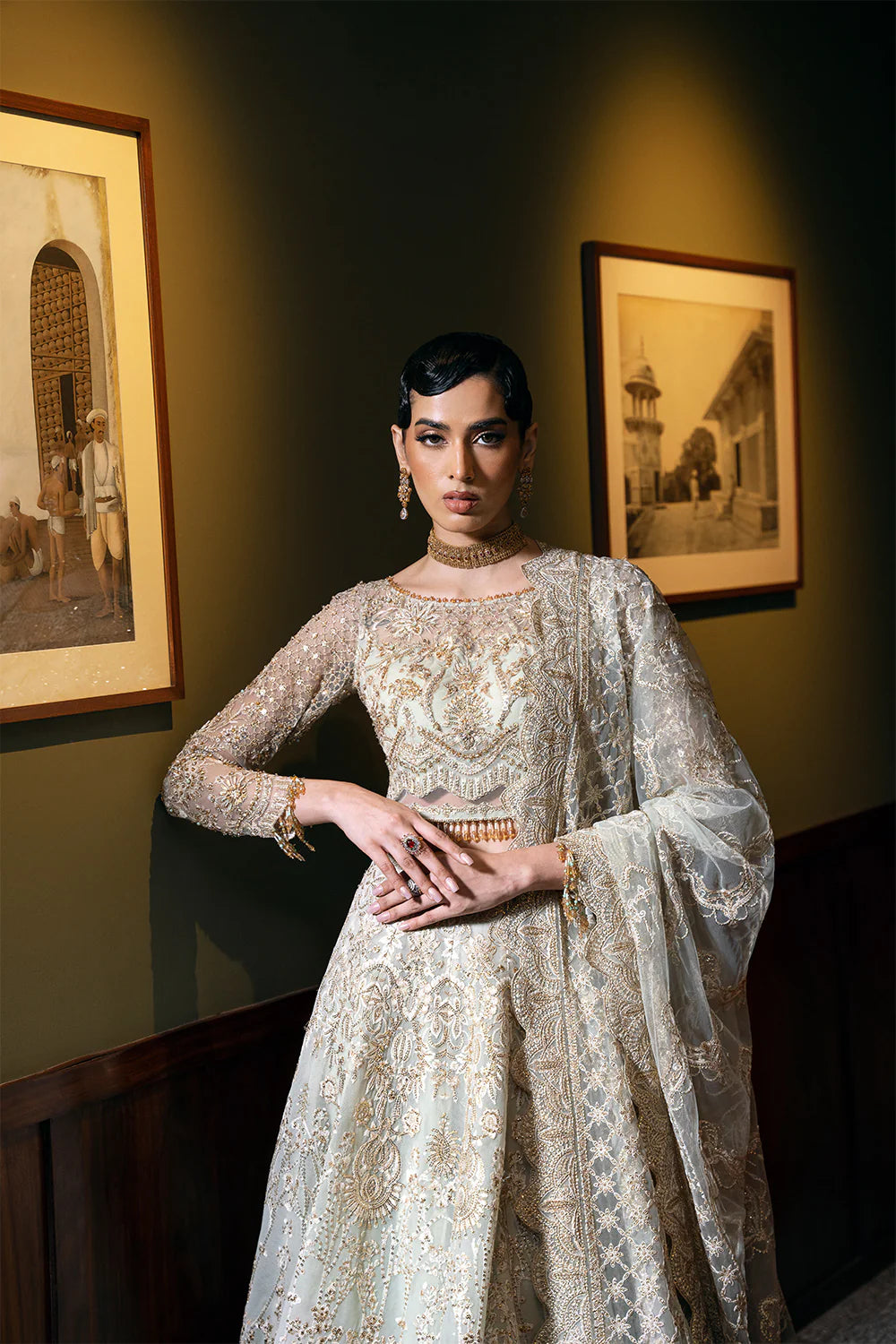 : Front view of woman in Saira Rizwan Chinese Silver bridal dress with intricate embroidery, traditional choker necklace, and elegant pose in sophisticated interior with vintage artwork