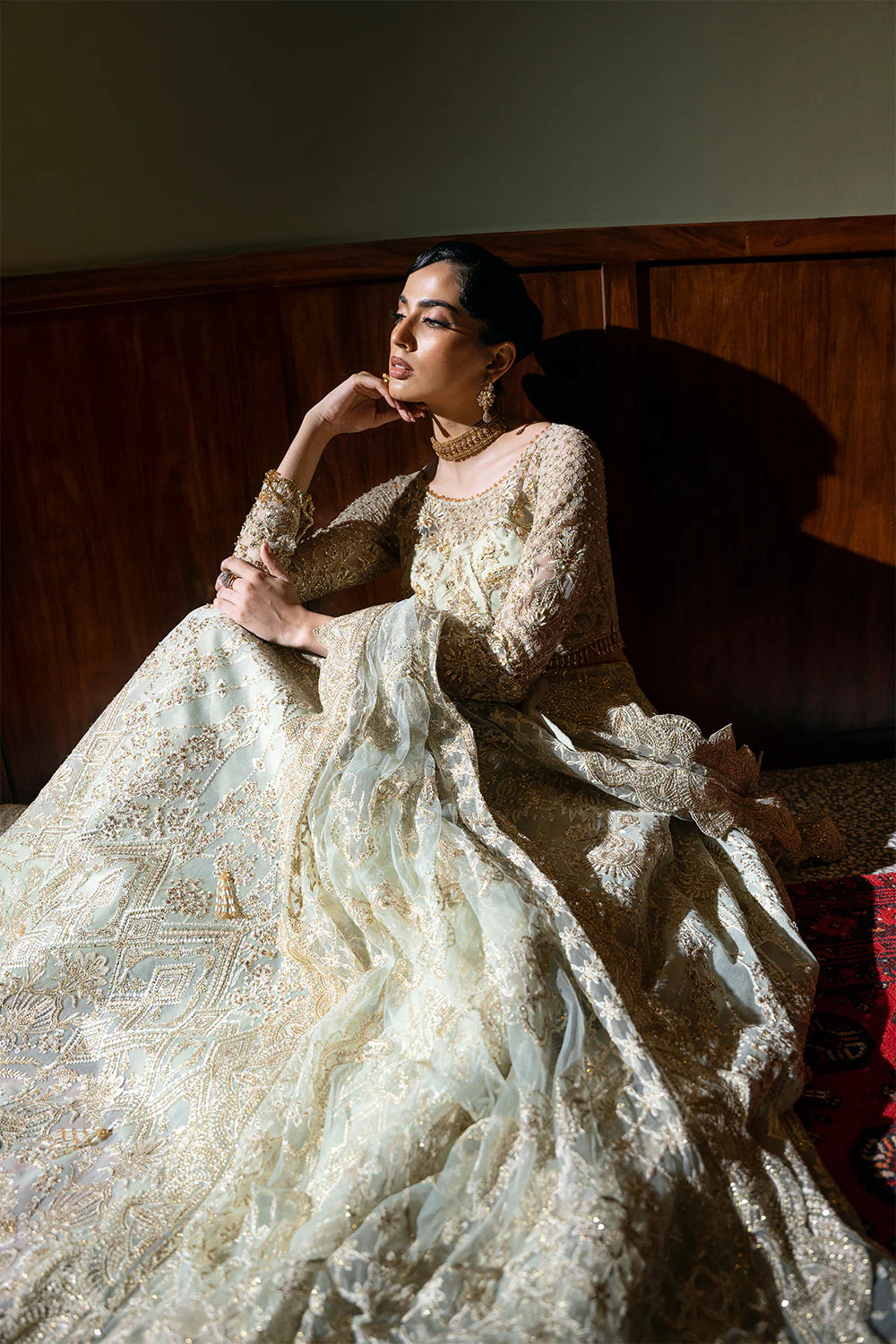 Relaxed seated pose of woman in Saira Rizwan Chinese Silver bridal lehenga with intricate embroidery, showcasing the flowing fabric and elegant traditional styling against wood paneled interior