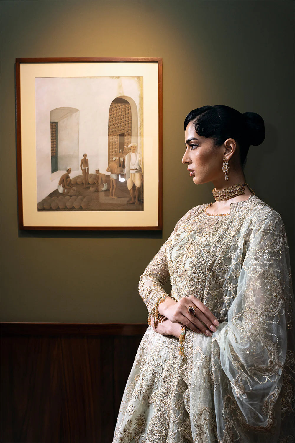 Side profile of Pakistani bridal outfit showcasing detailed embroidery work on net fabric, traditional gold jewelry, and elegant styling against vintage framed artwork backdrop