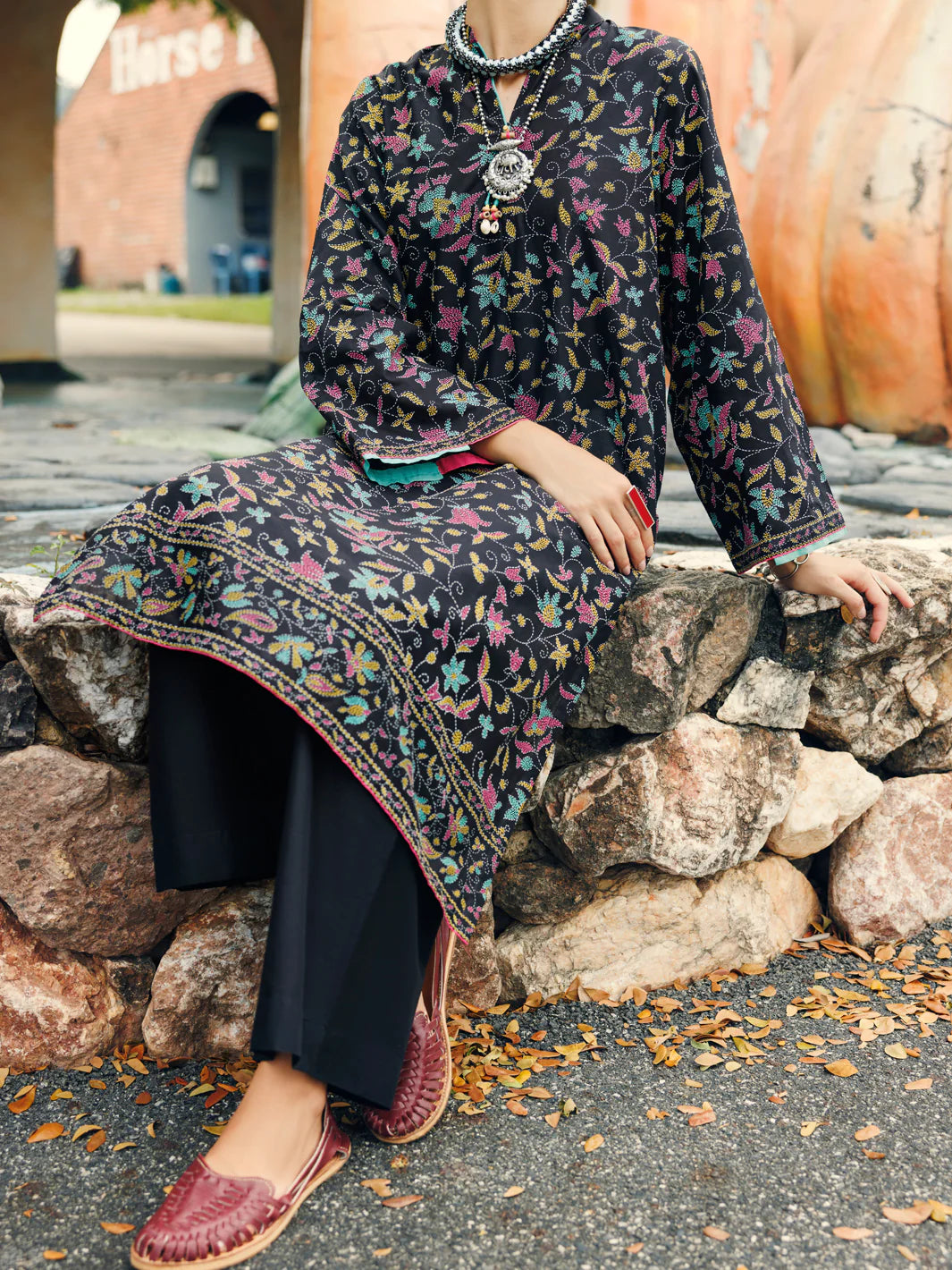 Woman sitting rocks 3-quarter front view black floral lawn kurti embroidered border hem Almirah Eid festive Pakistani suits online Canada