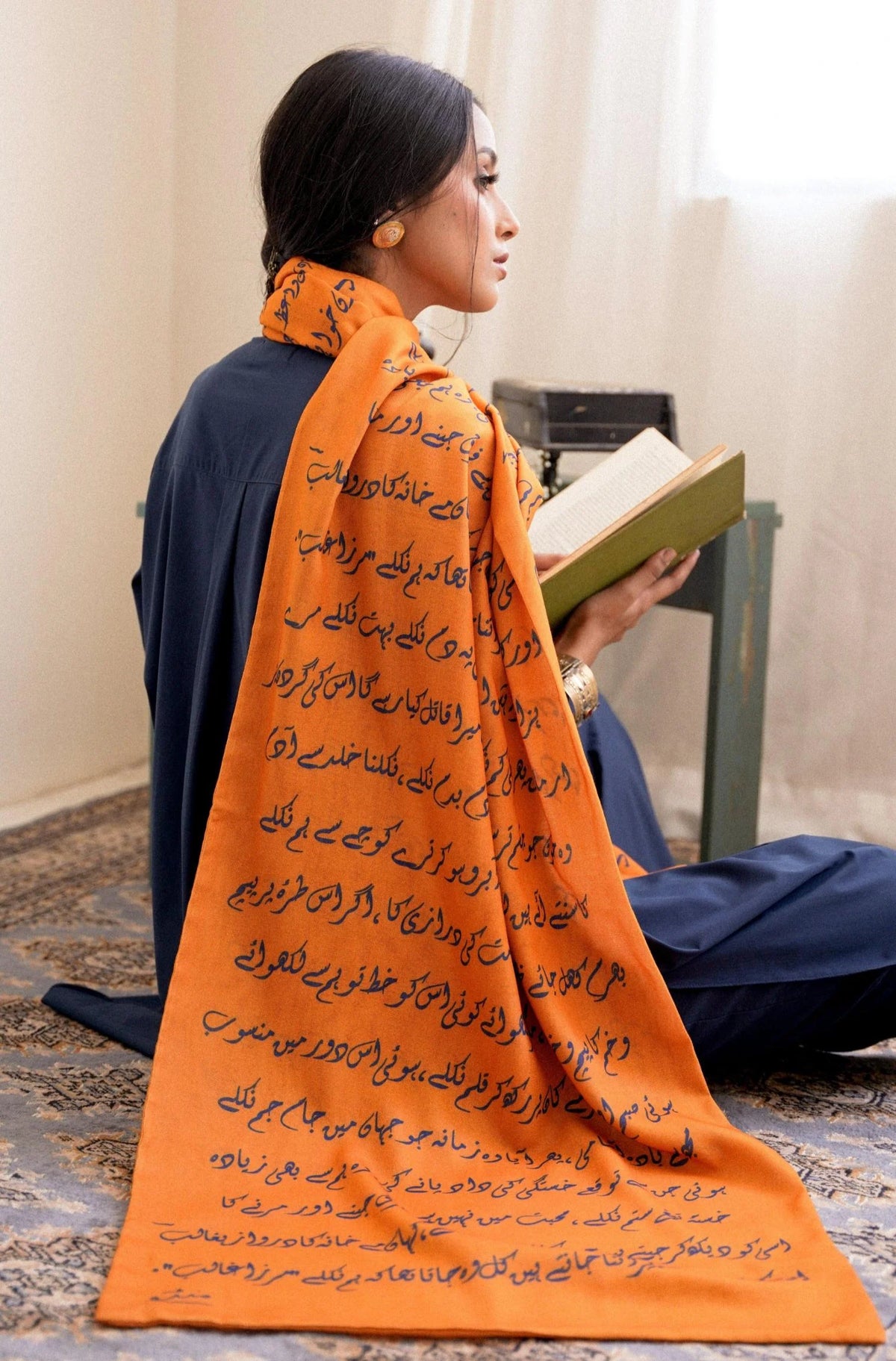 Woman in contemplative pose reading book while wearing orange Armaan stole with Urdu poetry print, showcasing the literary inspiration behind Manto's design