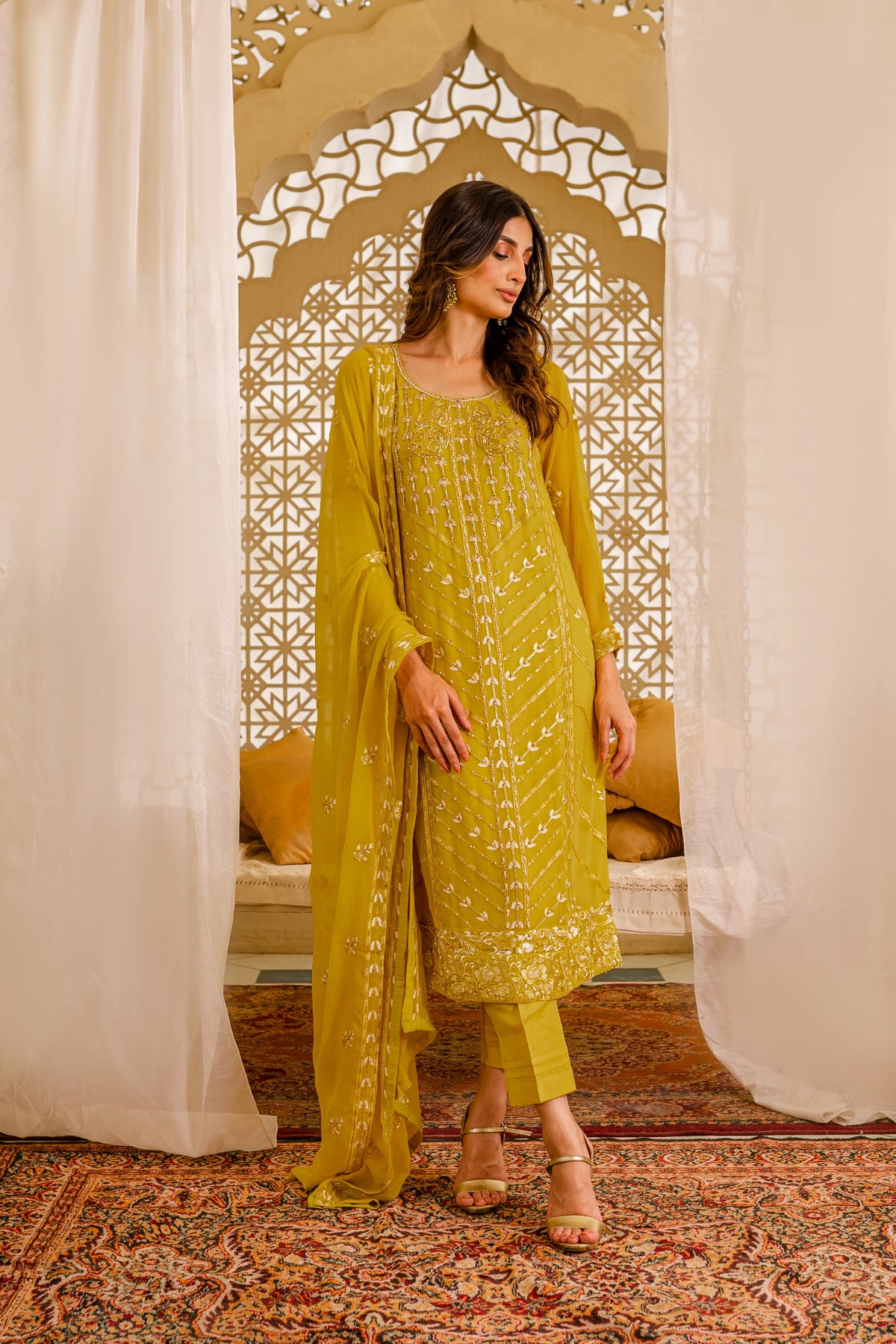 A woman standing in a traditional, ornate room wearing a mustard yellow chiffon outfit with hand embellishments and raw silk pants.