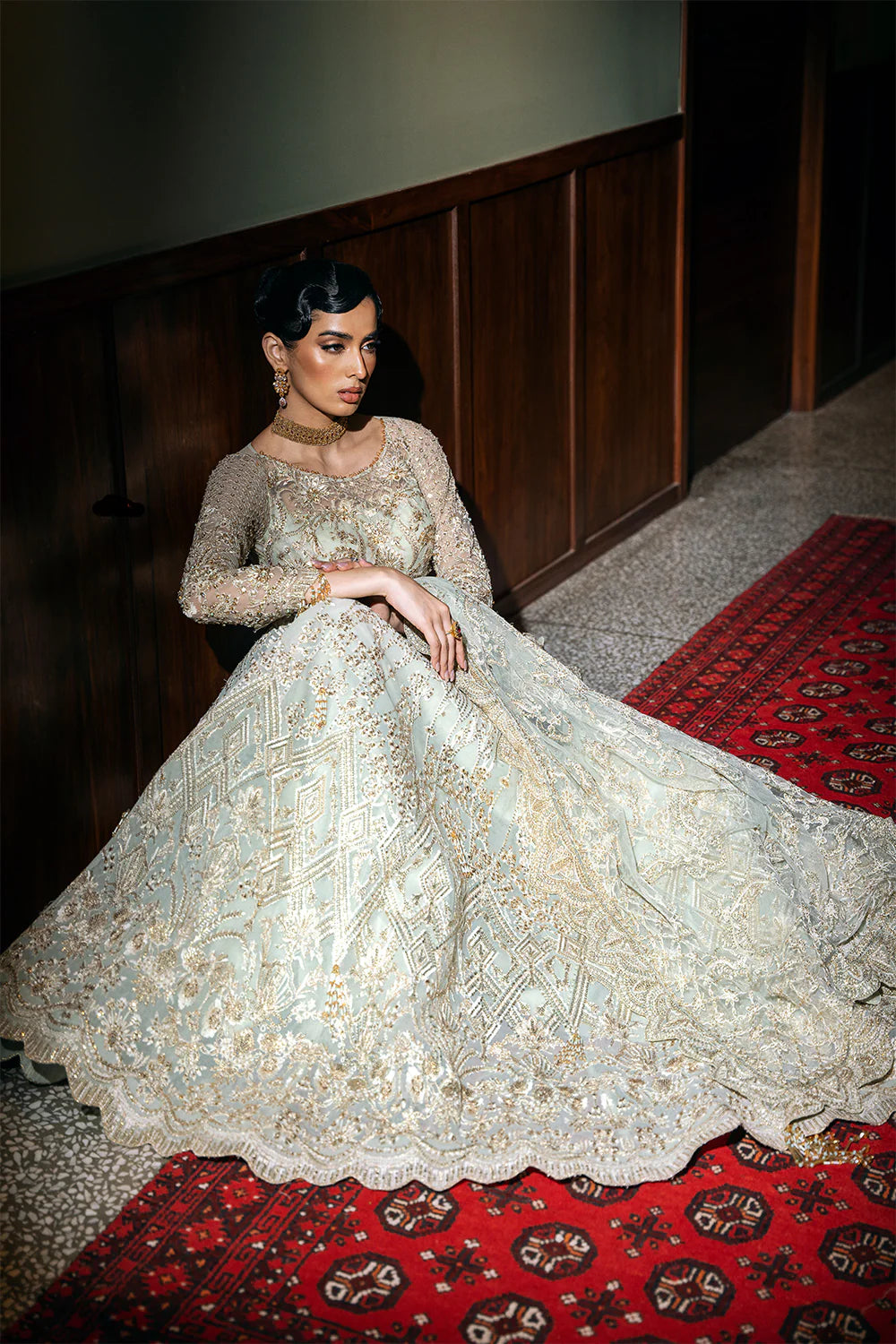 Seated pose displaying full spread of Pakistani bridal lehenga with Chinese Silver embroidery, showcasing the voluminous silhouette and intricate beadwork on traditional carpet