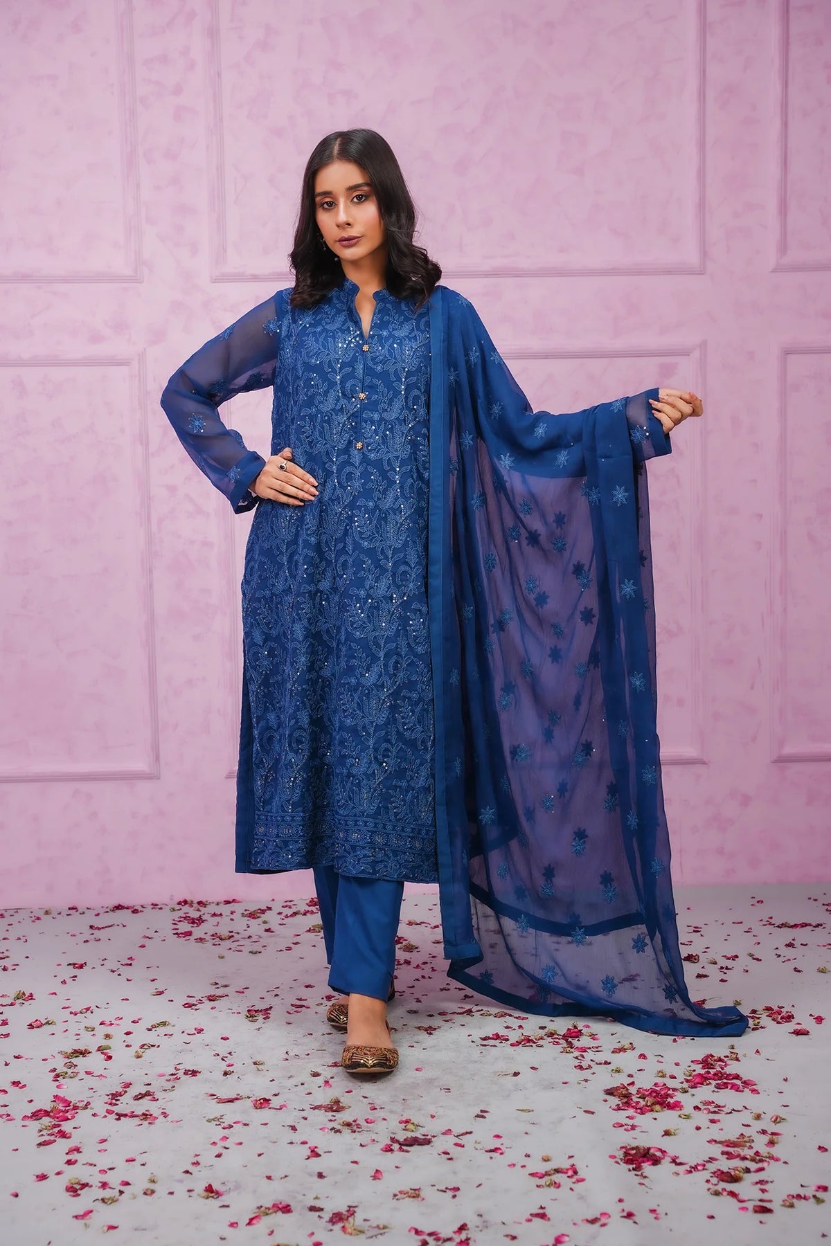 A woman posing in a blue chiffon stitched 3-piece suit with embroidery, including a shirt, dupatta, and trousers, standing on a background with pink flowers scattered around.